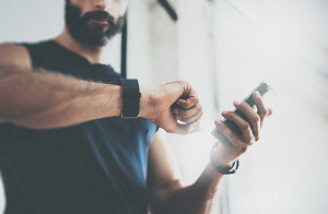 Close-up Shot Bearded Sportive Man After Workout Session Checks Fitness Results Smartphone.Adult Guy Wearing Sport Tracker Wristband Arm.Training hard inside gym.Horizontal bar background.Blurred.