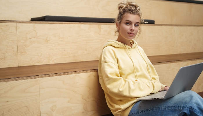 Female university student waiting for lecture, sitting with laptop on laps and looking camera. Woman freelancer working on laptop in co-working space. Girl sit in cafe on tribune, studying.
