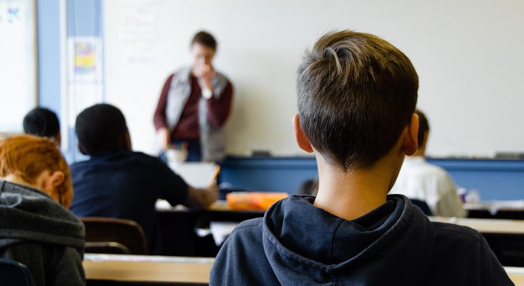 Joven alumno de espaldas en el aula con profesor al fondo. | Taylor Wilcox vía Unsplash 